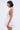 A woman with short dark hair stands in profile against a light background, modeling the SUMMER'S AROUND THE CORNER EYELET MINI DRESS—a sleeveless white halter-neck mini dress with a layered skirt, perfect for summer.