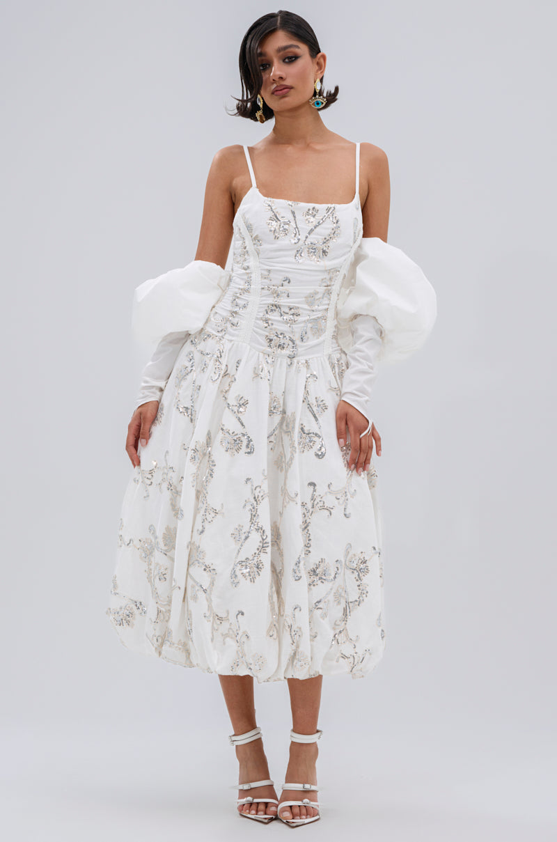 A woman wears the BEST DRESSED PRINTED MIDI DRESS, featuring floral embroidery, puffed long sleeves, thin straps, and a ruched bodice. She pairs it with white strappy heels and statement earrings against a plain background.