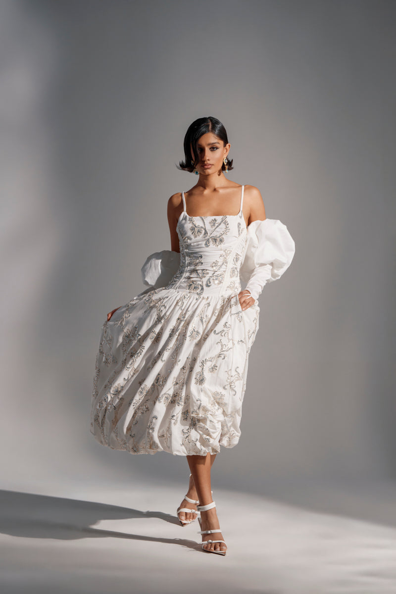 A woman models the BEST DRESSED PRINTED MIDI DRESS—a white midi with floral embroidery, thin straps, a single puff sleeve, and a bubble hem—paired with strappy heels and statement earrings against a softly lit backdrop.