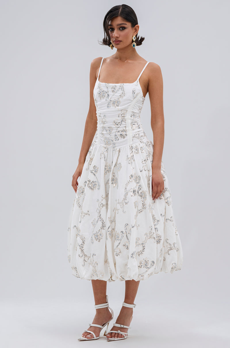 A woman models the BEST DRESSED PRINTED MIDI DRESS—an ankle-length white dress with thin straps and floral embroidery—styled with white strappy heels and statement earrings, set against a plain light gray background.