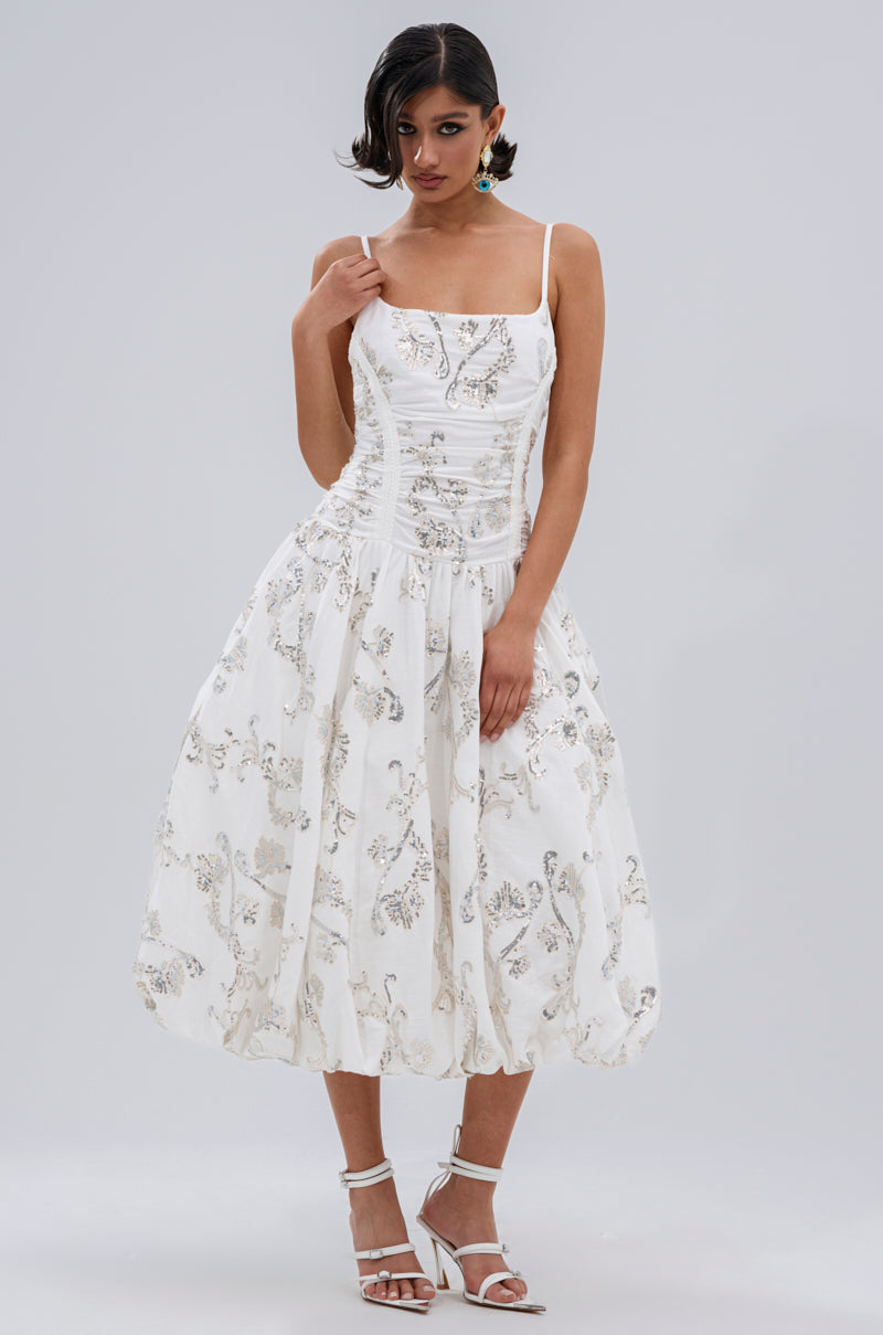 A woman with short dark hair wears the BEST DRESSED PRINTED MIDI DRESS, featuring silver floral embroidery, a ruched bodice, and flared skirt. She pairs it with statement earrings and white strappy heels against a plain background.
