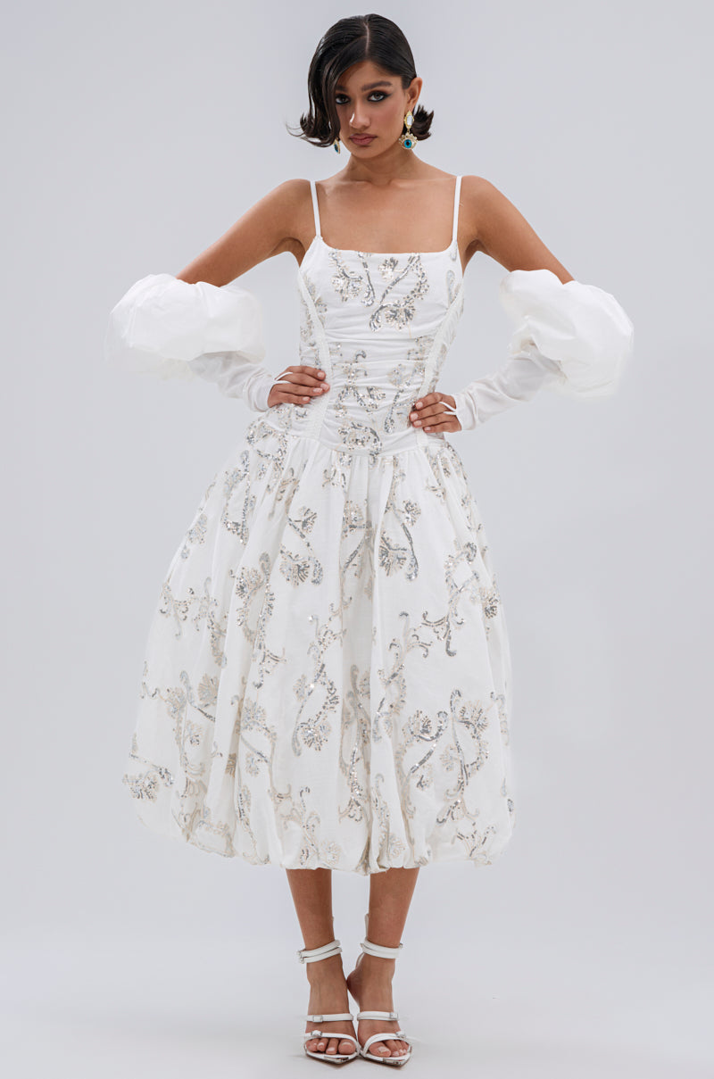 A woman models the BEST DRESSED PRINTED MIDI DRESS, featuring thin straps, floral embroidery, and puffed tulle sleeves. She pairs it with white strappy heels and has short, wavy hair styled to one side against a plain background.