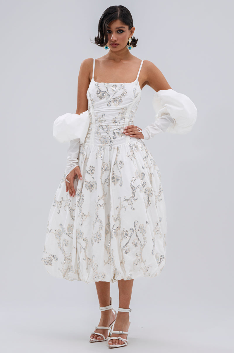 A woman wearing the BEST DRESSED PRINTED MIDI DRESS with silver embellishments, floral embroidery, thin straps, voluminous detachable sleeves, and white strappy heels stands against a plain light background.