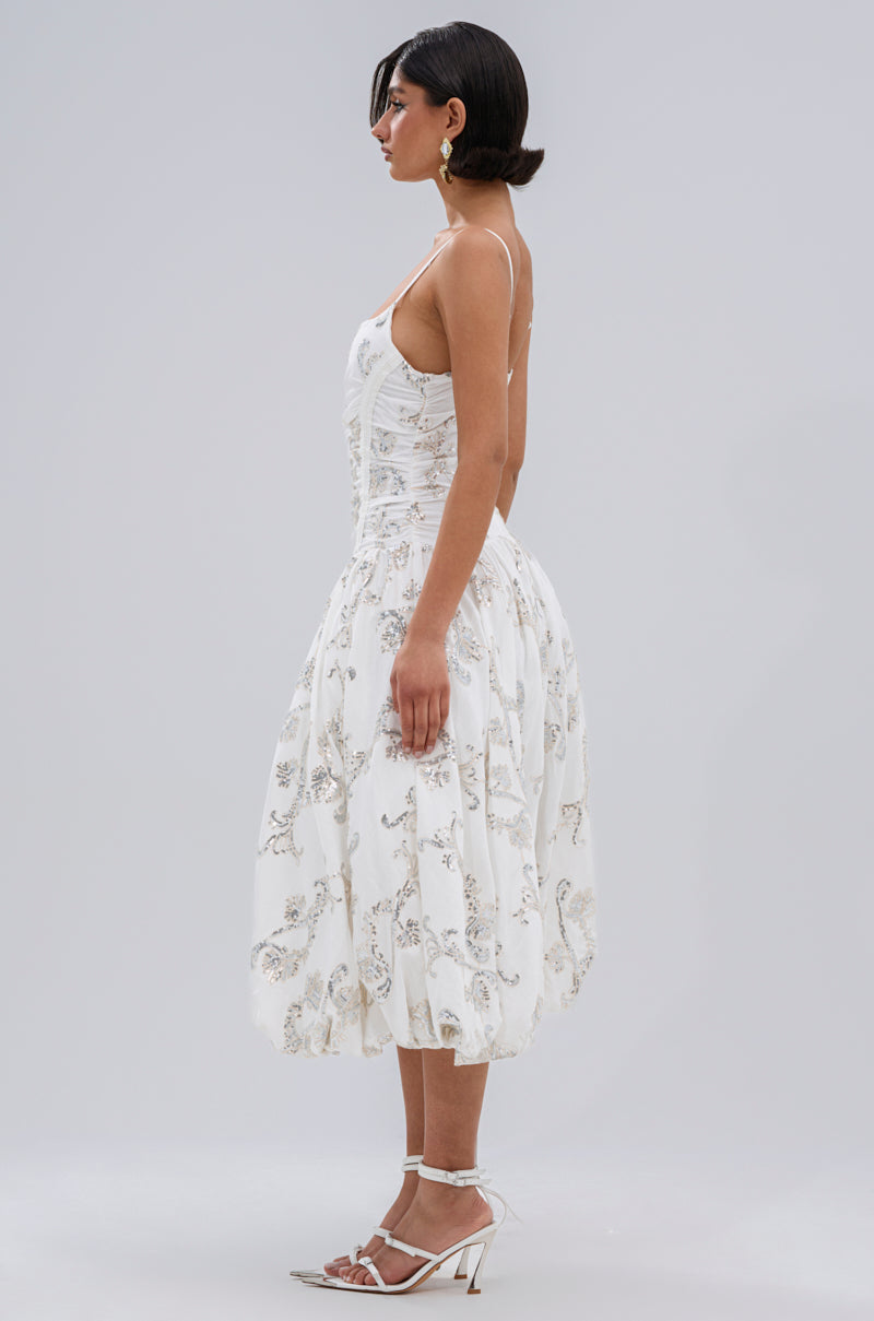 A woman with short dark hair stands in profile wearing the BEST DRESSED PRINTED MIDI DRESS, a white sleeveless midi with silver floral embroidery, paired with white high-heeled sandals against a plain light gray background.