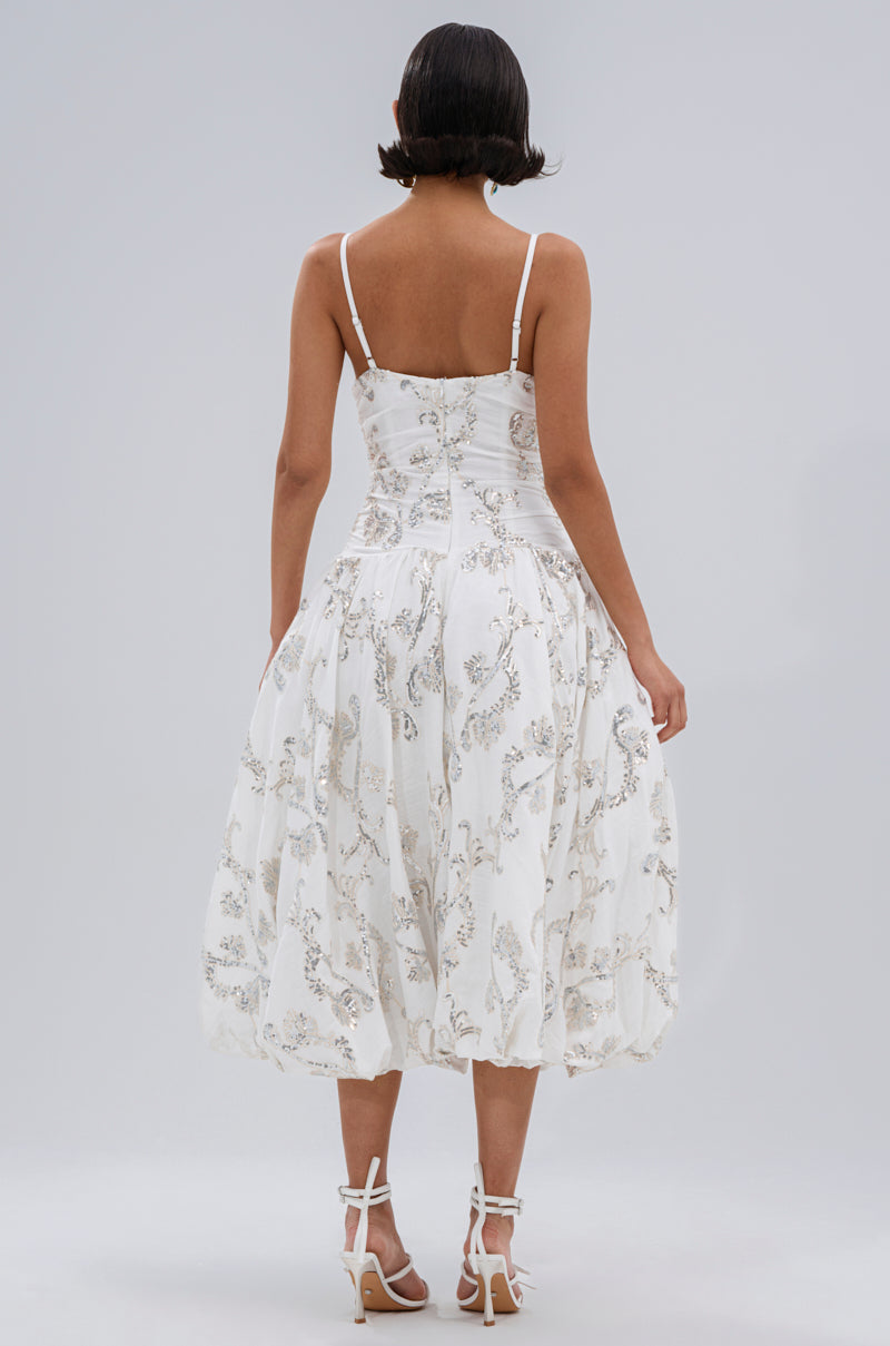 A woman with short dark hair models the BEST DRESSED PRINTED MIDI DRESS, featuring floral embroidery and thin straps. She stands with her back to the camera in white strappy heels against a plain gray background.