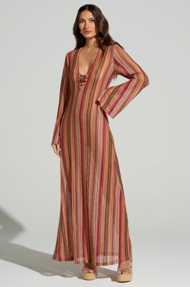 Against a plain backdrop, a person models the IBIZA DREAMIN CROCHET MAXI DRESS featuring stripes in pink, orange, and brown hues. The deep V-neck and long sleeves bring a perfect summer vibe to the ensemble, which is completed with platform sandals.