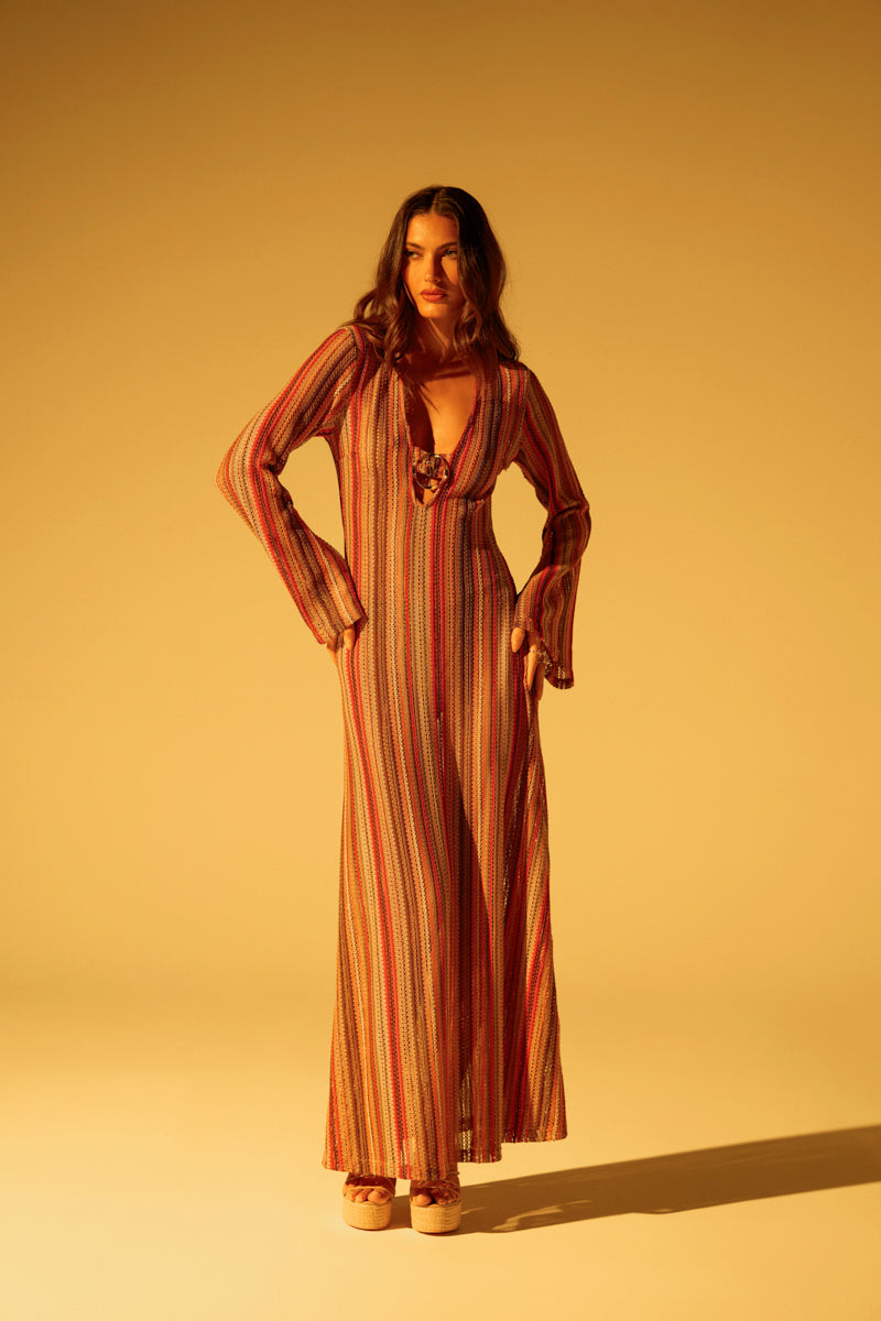 In a warmly lit room, a woman with long hair confidently wears the IBIZA DREAMIN CROCHET MAXI DRESS—featuring bell sleeves, a deep V-neckline, and multicolored stripes—exuding summer vibes as she looks at the camera.
