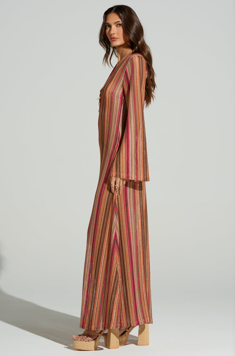 A woman with long brown hair effortlessly captures a summer vibe, wearing the IBIZA DREAMIN CROCHET MAXI DRESS with flared sleeves. She stands in profile, facing left, paired perfectly with platform wedge sandals against a plain, light-colored background.