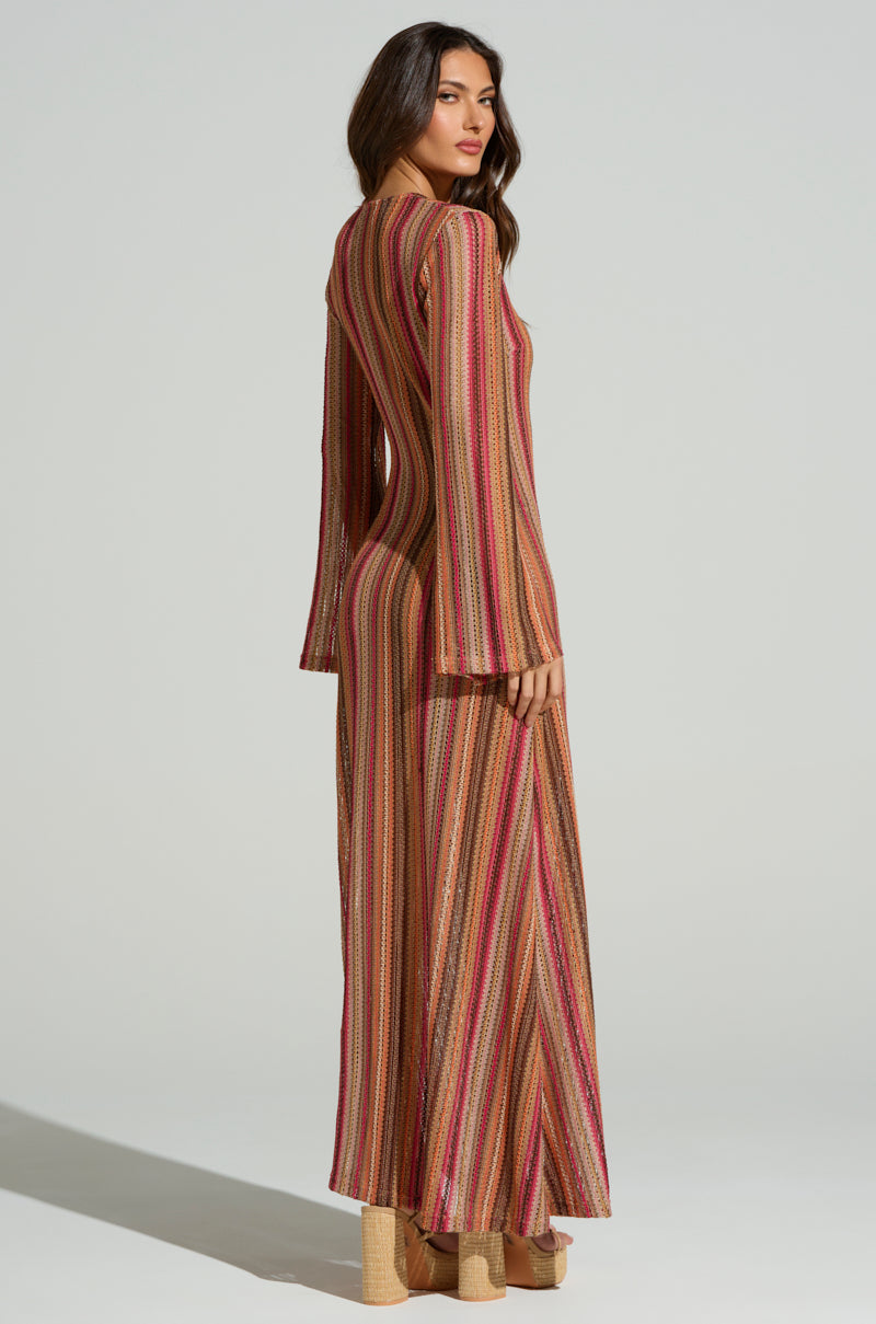 A person in the IBIZA DREAMIN CROCHET MAXI DRESS, featuring colorful stripes and bell sleeves, poses slightly back. Their long, wavy hair enhances the summer vibe as they pair it with platform shoes against a light neutral background.