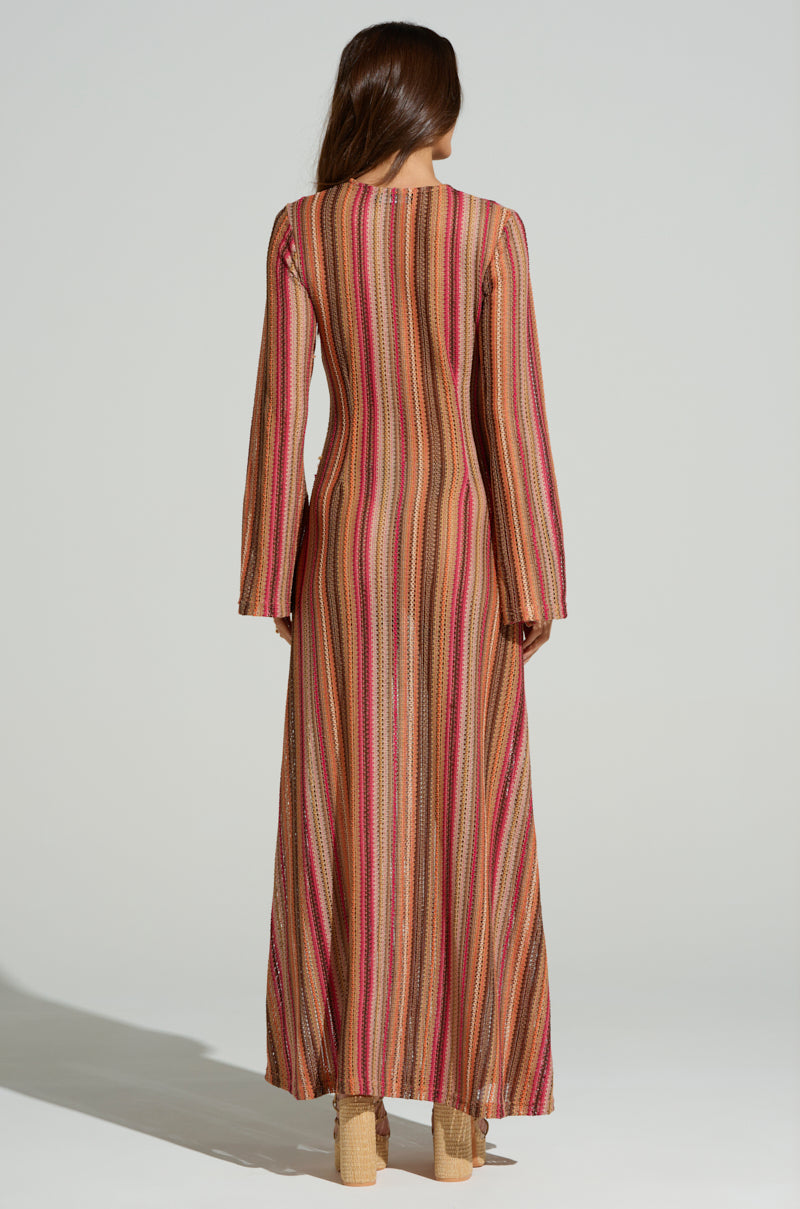 A person with long brown hair stands on a light background showcasing a summer vibe, wearing the IBIZA DREAMIN CROCHET MAXI DRESS, featuring colorful vertical stripes in red, pink, and beige. They have heeled sandals complementing the outfit.