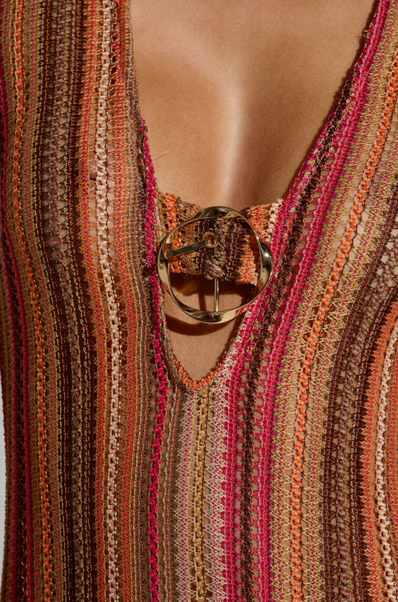 A person wears the IBIZA DREAMIN CROCHET MAXI DRESS, featuring vertical stripes in brown, orange, and pink. The V-neckline and large gold clasp on this knitted garment exude a summer vibe perfect for beachy days.