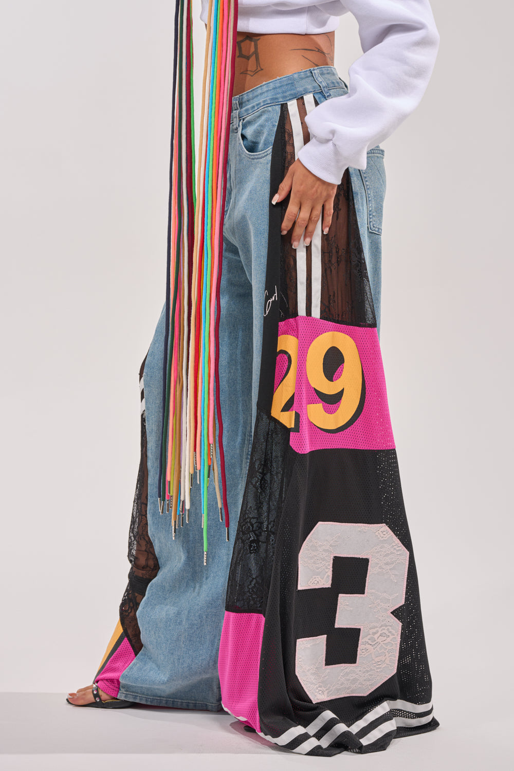 Someone in streetwear wears the GAME TIME RELAXED DENIM, a white cropped sweatshirt, and a vibrant patchwork skirt with large yellow "29," white and pink "3," and long multicolored strings hanging from the waist. 3