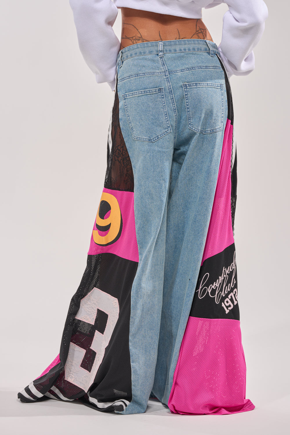 A person, back to the camera, wears GAME TIME RELAXED DENIM: streetwear-inspired wide-leg jeans with open sides that reveal pink, black, and white mesh panels featuring numbers and graphic text. 4