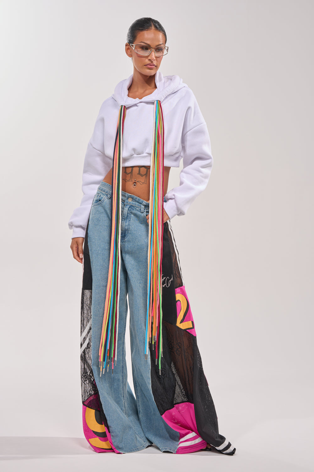 A woman poses in GAME TIME RELAXED DENIM jeans, a white cropped hoodie with multicolored fringe, glasses, and hoop earrings. Her hair is tied back, highlighting her vibrant streetwear style against a light gray background. 5