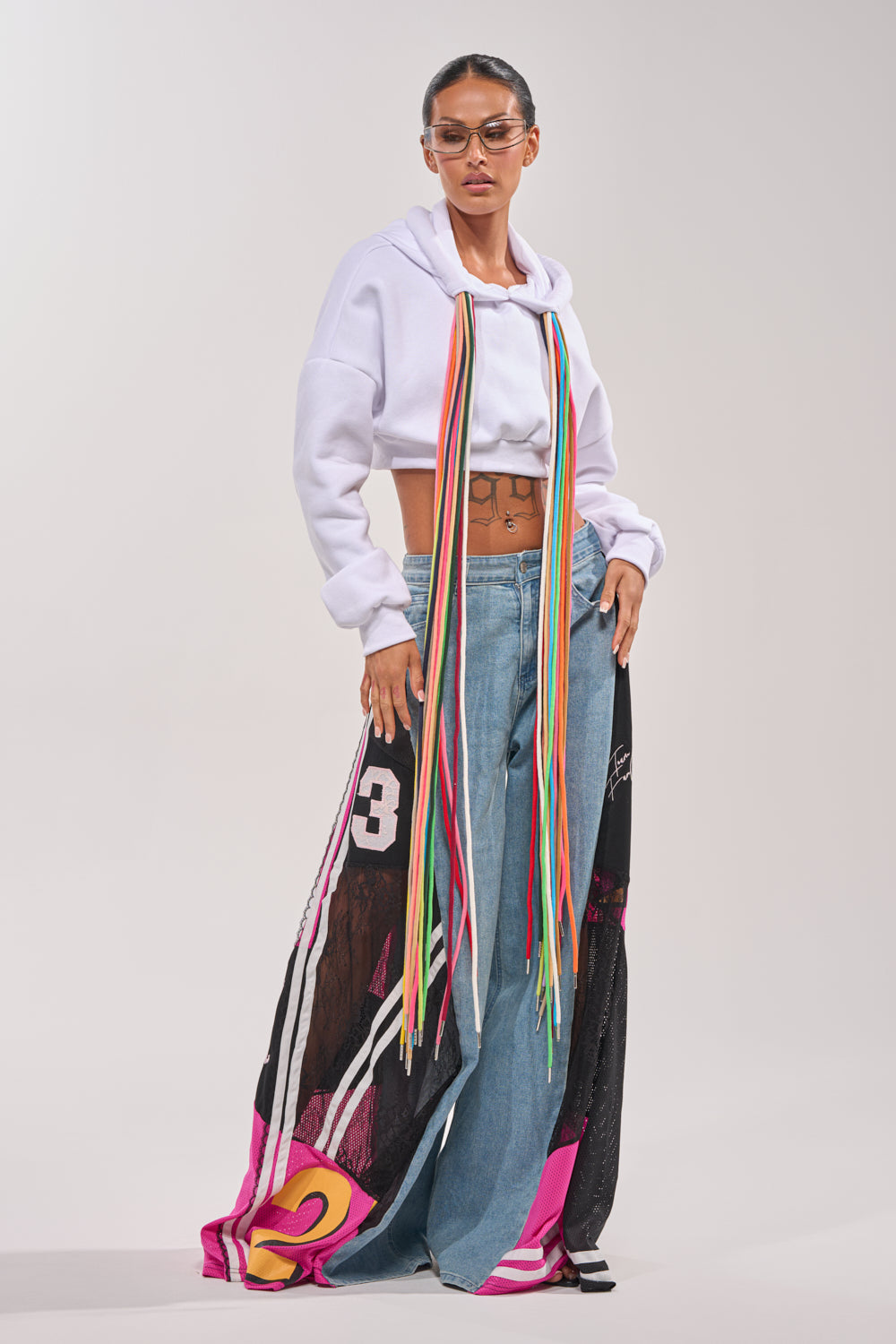 Wearing the GAME TIME RELAXED DENIM—wide-leg jeans with sheer, graphic-printed overlays—a person pairs them with a cropped white hoodie featuring bright ribbons, sunglasses, and stands confidently in streetwear style against a plain background. 6