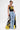 A woman stands confidently in high-rise DONT MESS WITH ME WIDE LEG DENIM PANT, a black tank top with "79", and a long yellow-black patchwork cape. She holds a small silver purse and sports short, styled hair.