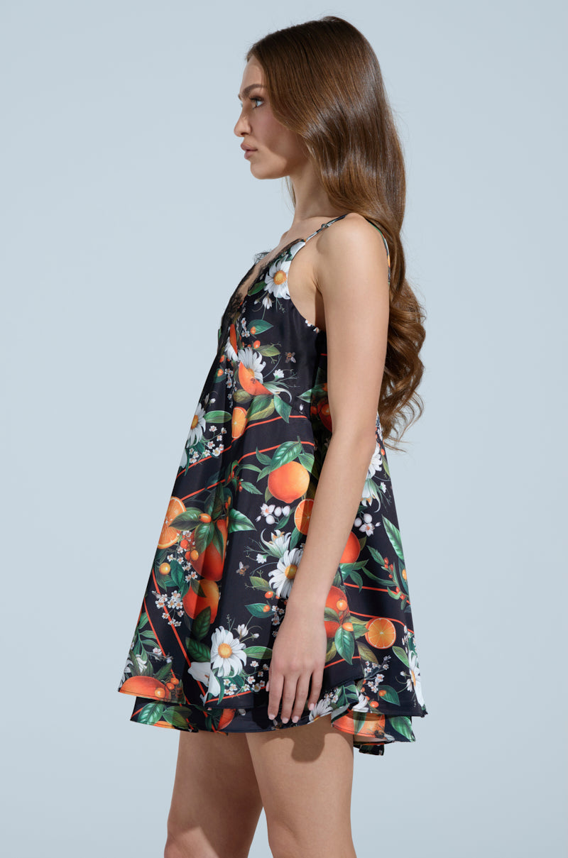 A woman with long, wavy brown hair stands in profile against a light blue background, wearing the TROPICAL GETAWAY SATIN MINI DRESS—a sleeveless black dress with a citrus and floral pattern, perfect for vacation.