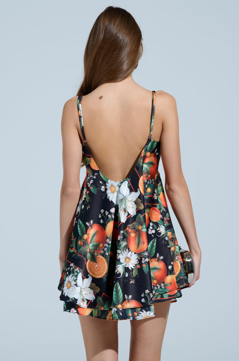 A woman with long brown hair faces away, wearing the TROPICAL GETAWAY SATIN MINI DRESS—a sleeveless, backless black mini dress with a bold citrus and floral print—holding a small clutch against a light blue background.