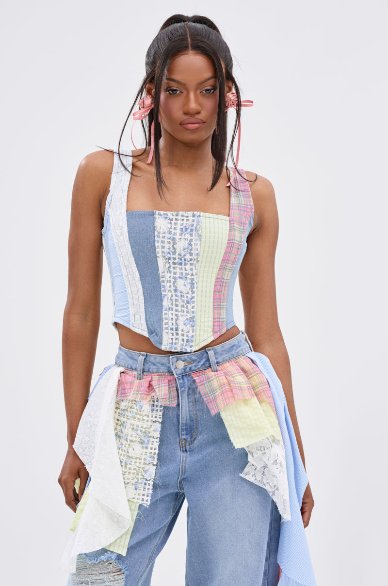A woman wears the LET THE FUN BEGIN MIXED PLAID CORSET TOP with blue jeans, standing against a light background. Her long straight hair is styled in pigtails tied with pink ribbons.