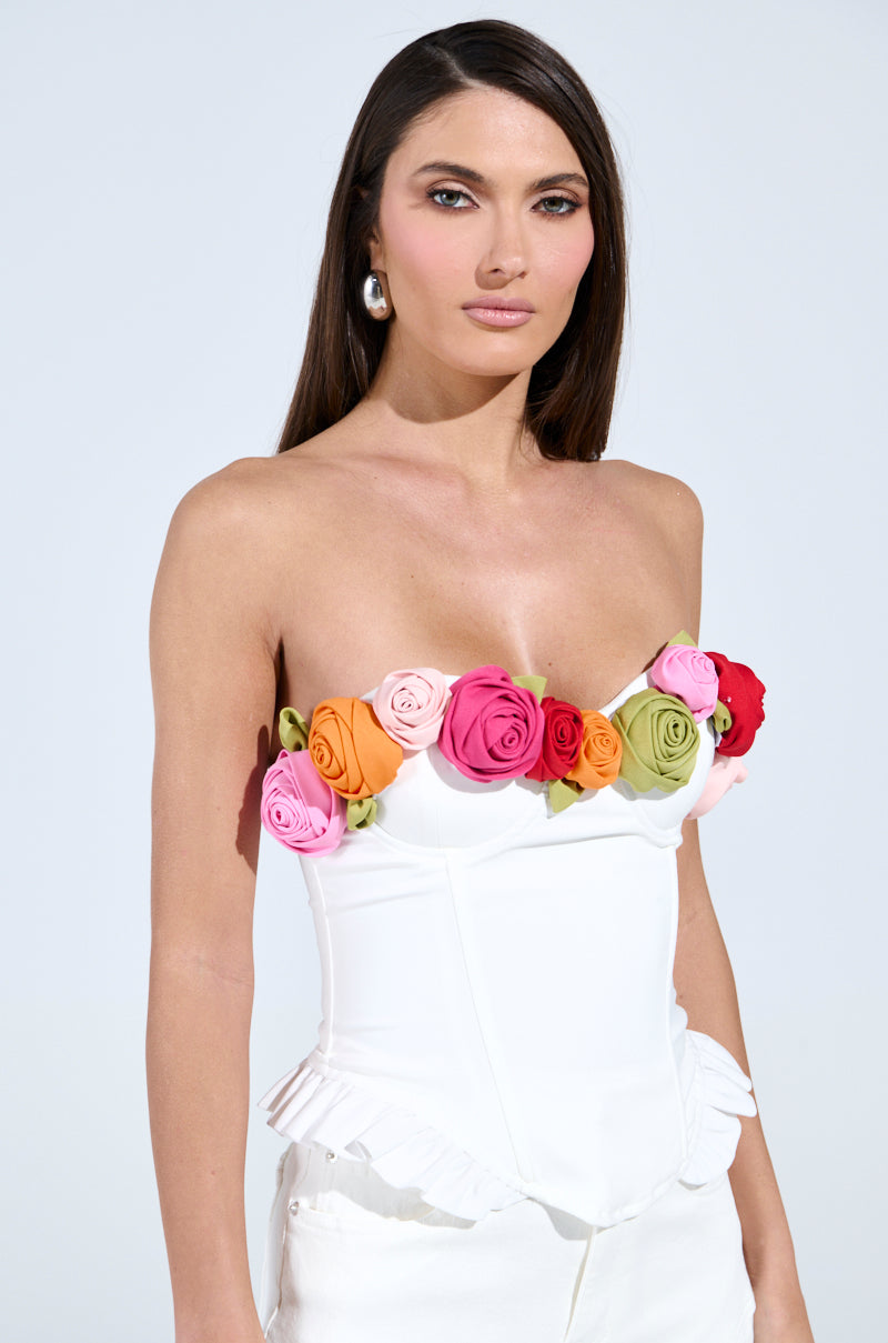 The woman dons the UNLEASH YOUR INNER DIVA FLOWER TRIM CORSET TOP, featuring a white strapless design with colorful fabric flowers at the neckline. Her long dark hair accents silver hoop earrings, and the adjustable lace-up back adds flair. The plain background is light grey.