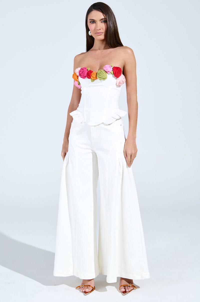 A woman poses against a plain background, wearing a white strapless jumpsuit featuring the "Unleash Your Inner Diva Flower Trim Corset Top" adorned with colorful roses. Her long brown hair complements the elegant adjustable lace-up back and her stylish brown strappy heels.