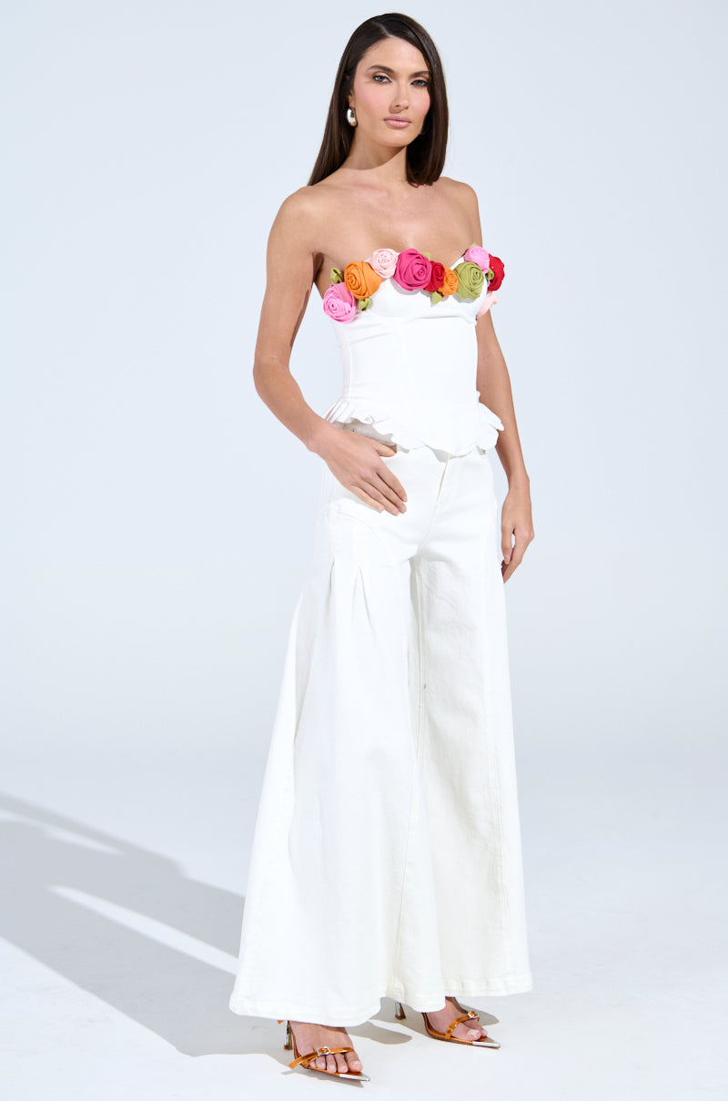 A woman confidently wears the "UNLEASH YOUR INNER DIVA FLOWER TRIM CORSET TOP," a strapless white top with colorful roses and flower trim. Paired with wide-legged white pants and high heels, her ensemble features an adjustable lace-up back, perfect for a stylish look in the bright studio setting.