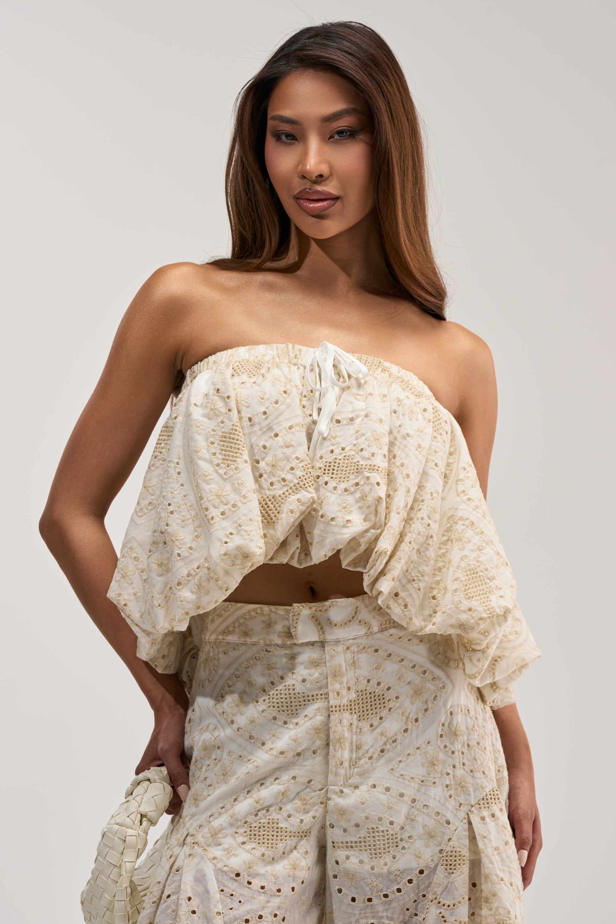 A woman wearing the SUMMER TIME BLISS EYELET TUBE TOP, a cream-colored, strapless summer top with ruffled details and a delicate eyelet pattern. She pairs it with matching high-waisted shorts, carries a small woven bag, and has long, straight hair.