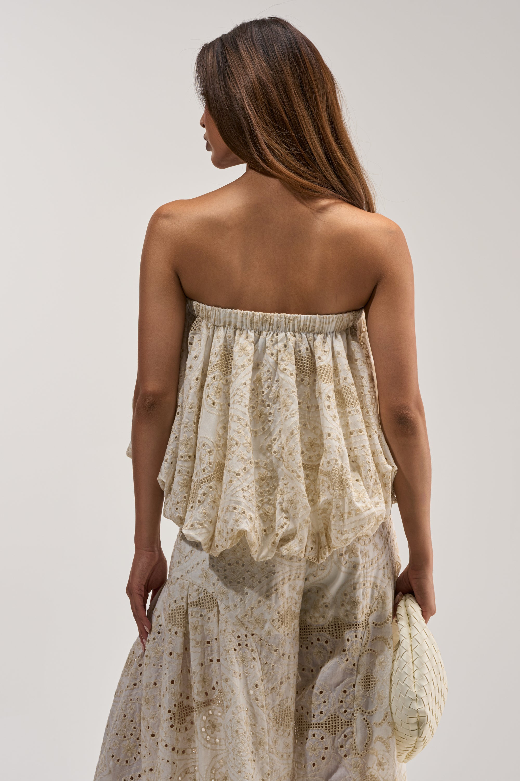 A woman with long brown hair wears the SUMMER TIME BLISS EYELET TUBE TOP, styled as a loose-fitting, cream-colored strapless dress, and holds a woven clutch. She is facing away from the camera against a plain background.