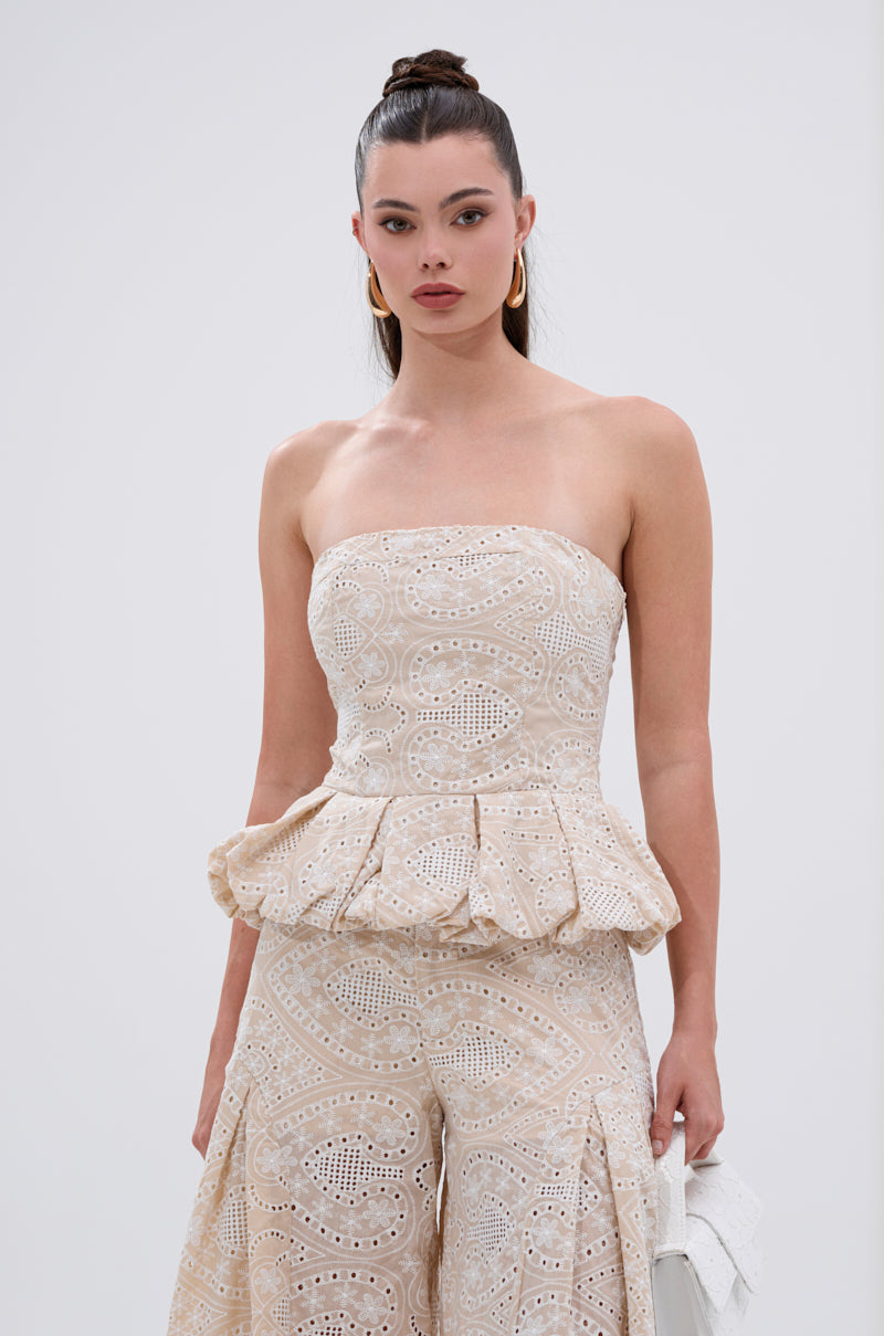 A woman with long dark hair in a high bun models the SUMMER TIME BLISS EYELET CORSET, showcasing its embroidered eyelet pattern and peplum hem, paired with matching wide-leg pants. She accessorizes with a white bag and large gold hoops. 0