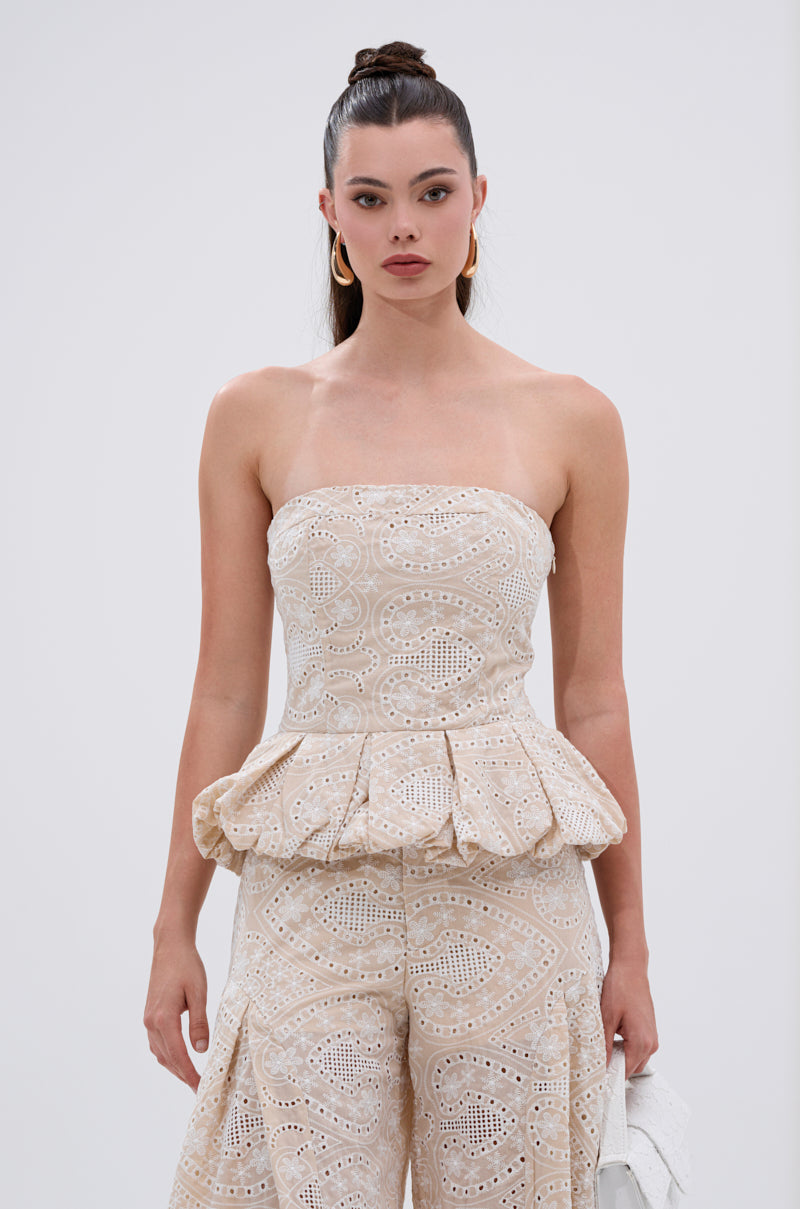 A woman with long dark hair in a high bun wears the SUMMER TIME BLISS EYELET CORSET, a strapless cream embroidered piece paired with matching wide-leg pants. She accessorizes with gold hoop earrings and holds a white bag against a plain white background. 1