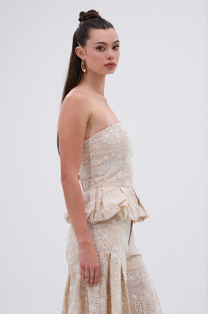 A woman with long dark hair styled half-up wears the SUMMER TIME BLISS EYELET CORSET, a strapless beige lace piece with embroidered eyelet and peplum details, standing sideways against a plain white background. 2