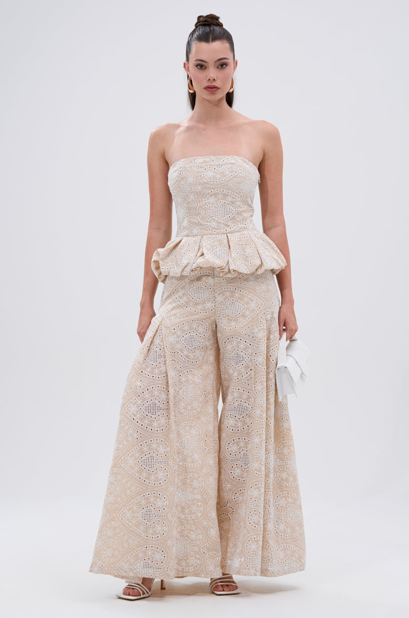 A woman stands against a plain background wearing the SUMMER TIME BLISS EYELET CORSET, paired with wide-leg pants. She accessorizes with a white clutch and strappy heels, her long hair tied back for an effortless look. 5