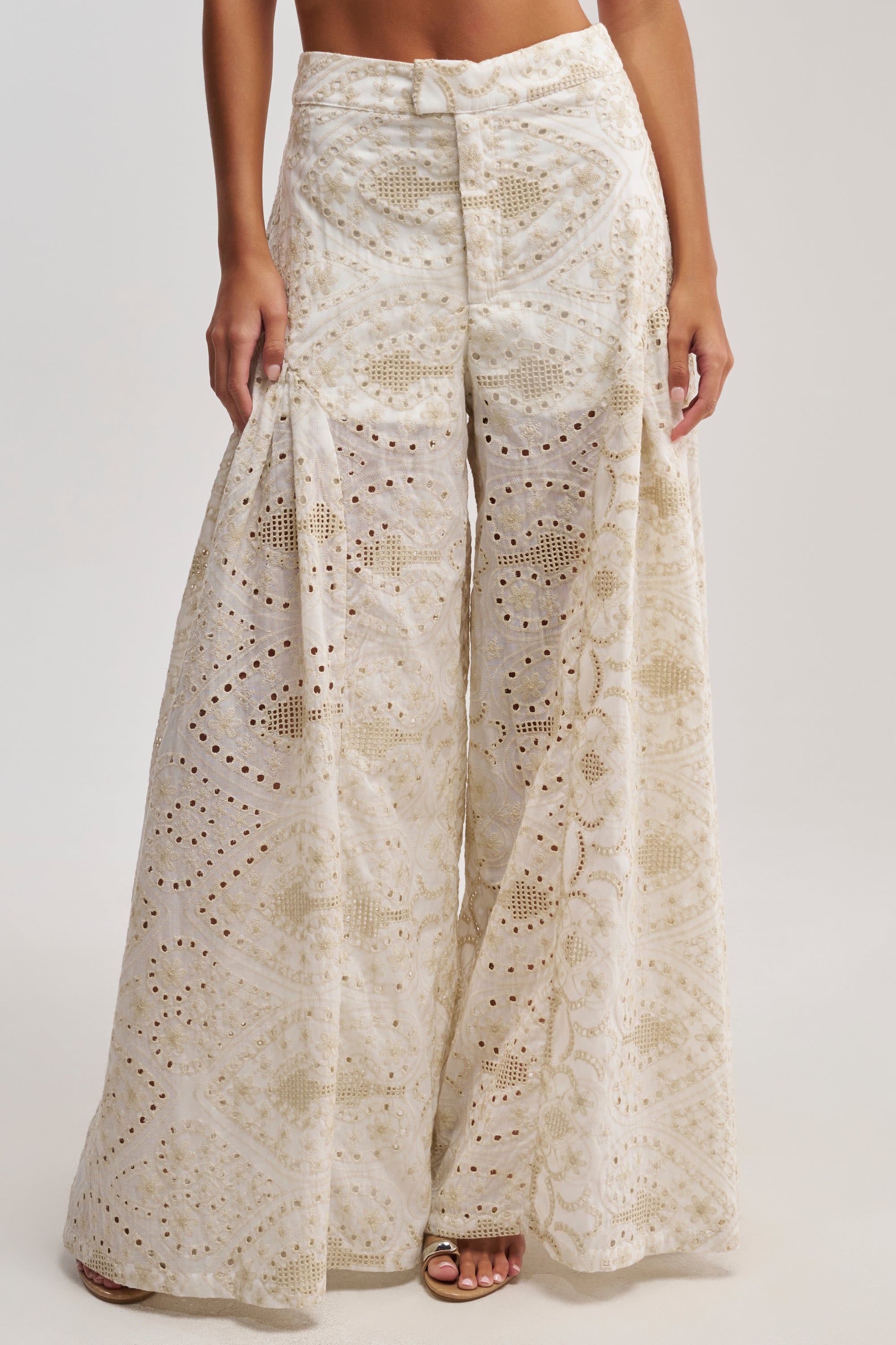 A woman wears the SUMMER TIME BLISS EYELET TROUSER, featuring high-waisted cream wide legs with intricate cutouts and geometric patterns, styled with open-toe shoes—ideal for chic, effortless vacation looks.