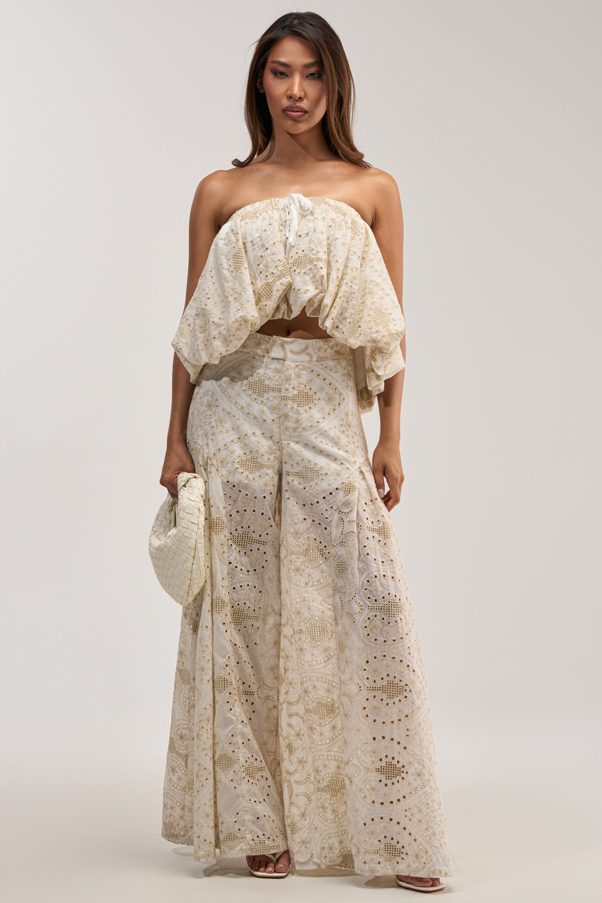 A woman with long, dark hair models the SUMMER TIME BLISS EYELET TROUSER—a cream-colored, wide-leg pant adorned with ivory lace and delicate eyelet details—paired with a matching cropped, ruffled strapless top. She completes the look with a white woven handbag against a plain, light gray studio backdrop.