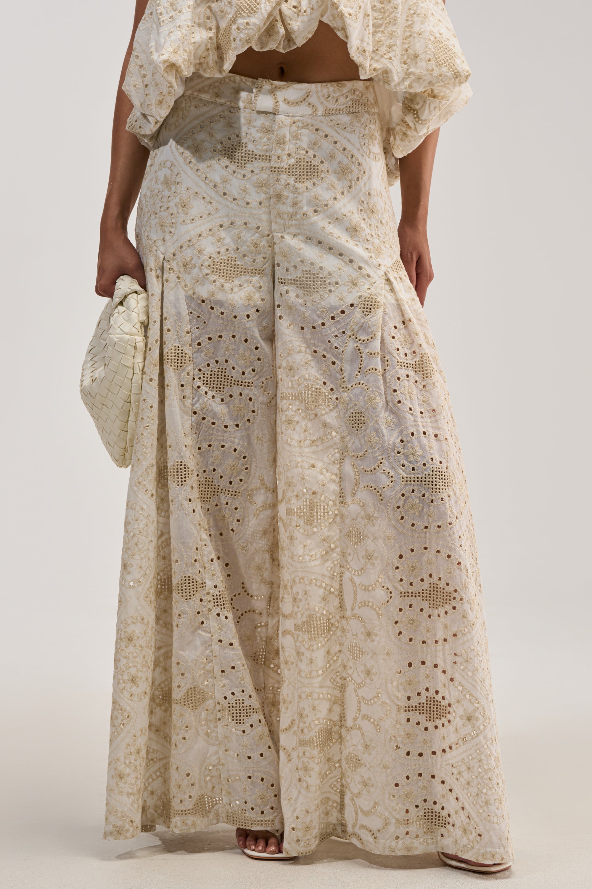 A woman is shown from the waist down wearing the SUMMER TIME BLISS EYELET TROUSER—ivory lace pants adorned with delicate floral embroidery—paired with a matching crop top and holding a woven beige clutch, set against a plain light background.