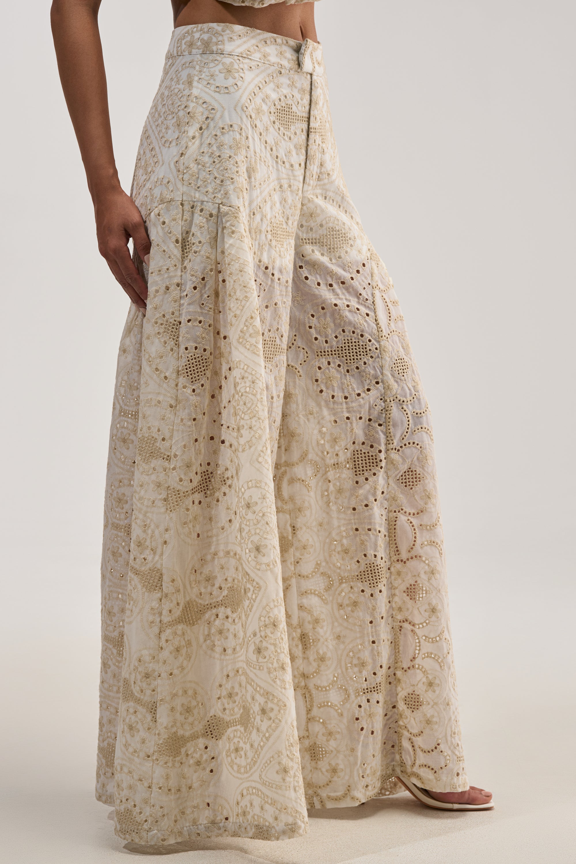 Dressed in the SUMMER TIME BLISS EYELET TROUSER, this individual showcases high-waisted, wide-leg trousers in a creamy ivory hue. The floor-grazing pants feature delicate eyelet lacework and subtle geometric patterns, creating a flowy silhouette that pairs beautifully with open-toe shoes.