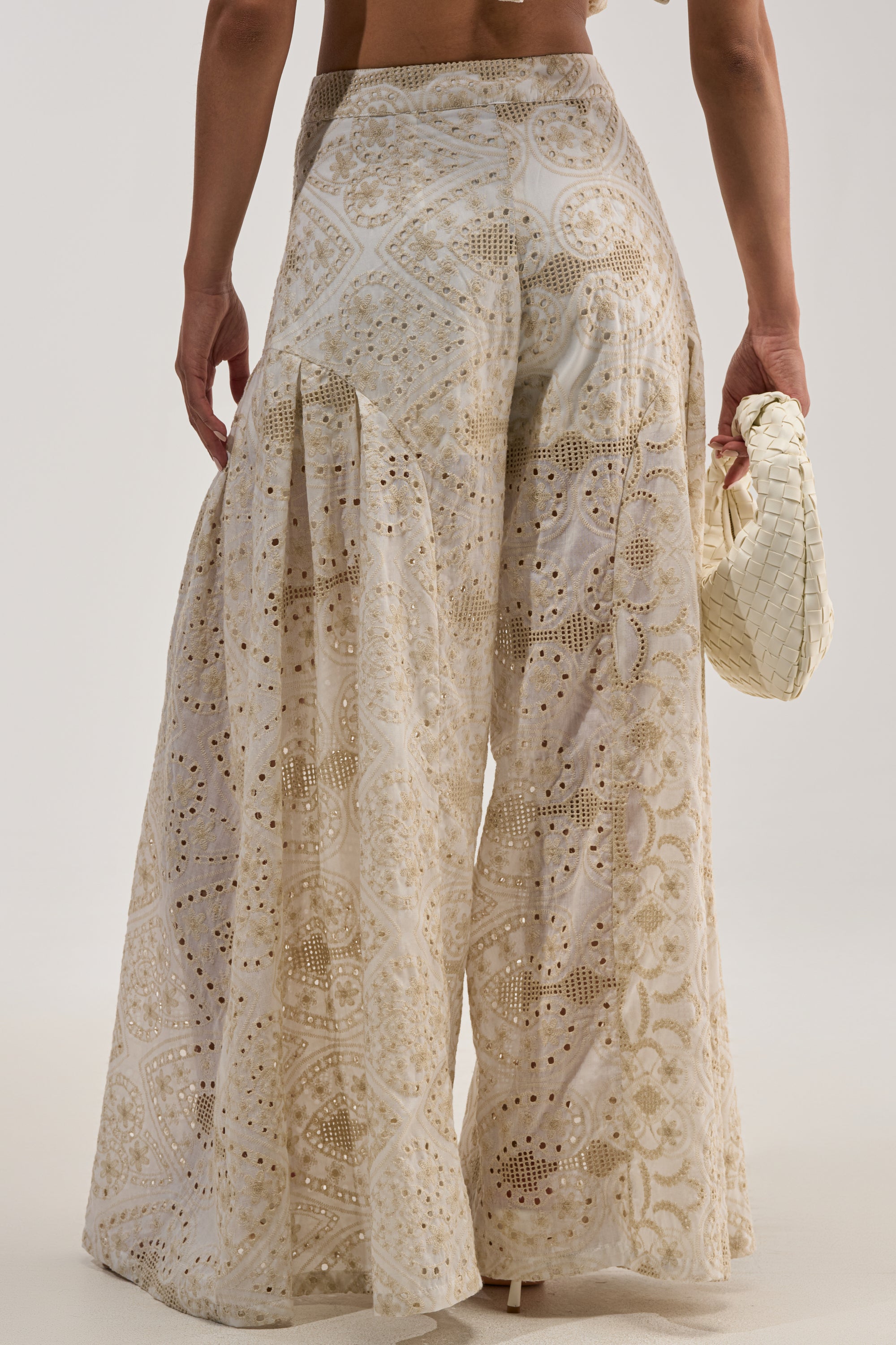 A person stands against a plain background wearing the SUMMER TIME BLISS EYELET TROUSER, which features intricate ivory lace patterns, and holds a woven beige clutch.