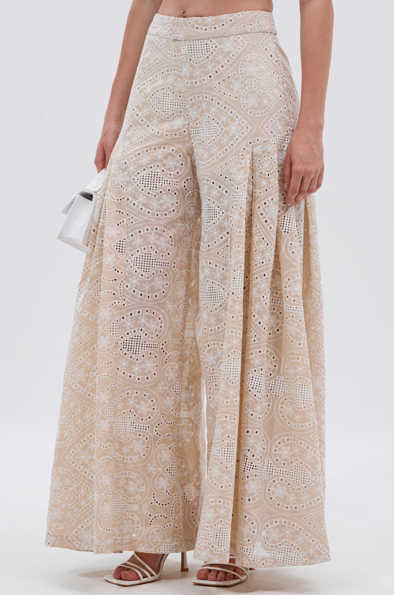 Someone in the SUMMER TIME BLISS EYELET TROUSER—high-waisted, wide-leg pants with an intricate cut-out design—paired with a small white handbag and strappy heeled sandals, ideal for summer days. 1