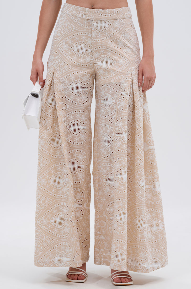 Wearing the SUMMER TIME BLISS EYELET TROUSER—wide-leg, high-waisted beige pants with detailed cut-outs—paired with white heeled sandals and a small white handbag, set against a plain white background. 2