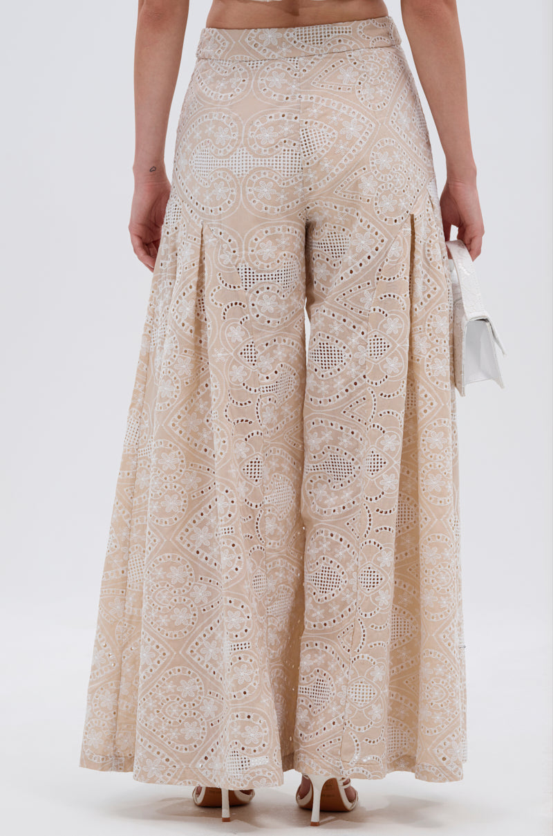 A woman radiates style in the SUMMER TIME BLISS EYELET TROUSER, featuring wide-leg beige fabric with intricate cutouts. Shown from the back, she pairs it with a small white purse and high heels. 4