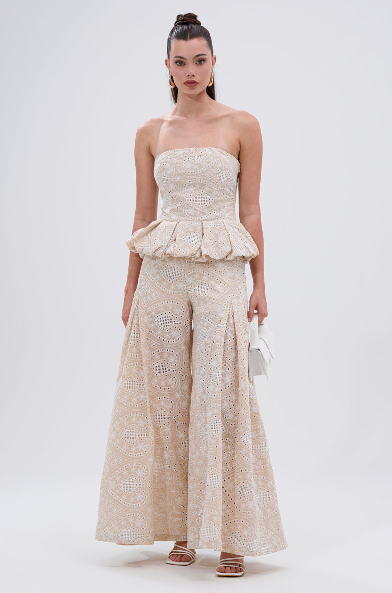 A woman poses against a plain background in the SUMMER TIME BLISS EYELET TROUSER set: a strapless cream peplum top and high-rise, wide-leg pants with an embroidered eyelet pattern, styled with a white clutch and strappy heels. 5