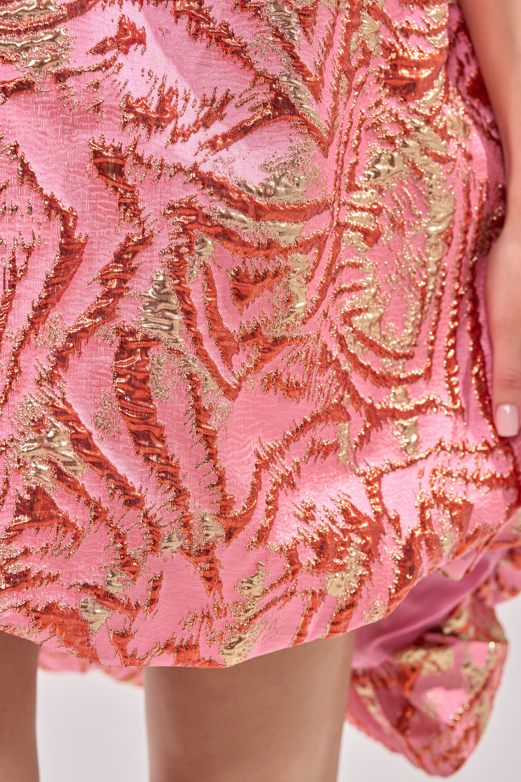 A close-up of the LOVER GIRL BROCADE BUBBLE DRESS IN PINK shows its metallic strapless design with gold and red textured patterns as a hand with light pink nail polish rests at the edge. 9