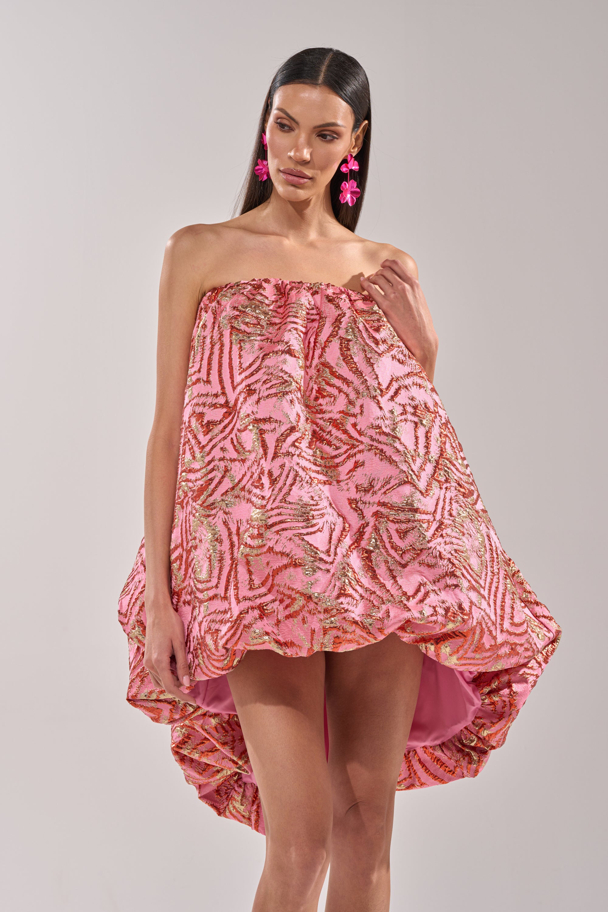 A woman wears the LOVER GIRL BROCADE BUBBLE DRESS IN PINK, a strapless mini dress with a high-low hem and pink-gold sequin patterns. She has straight dark hair and bold pink flower-shaped earrings, standing against a plain background. 1