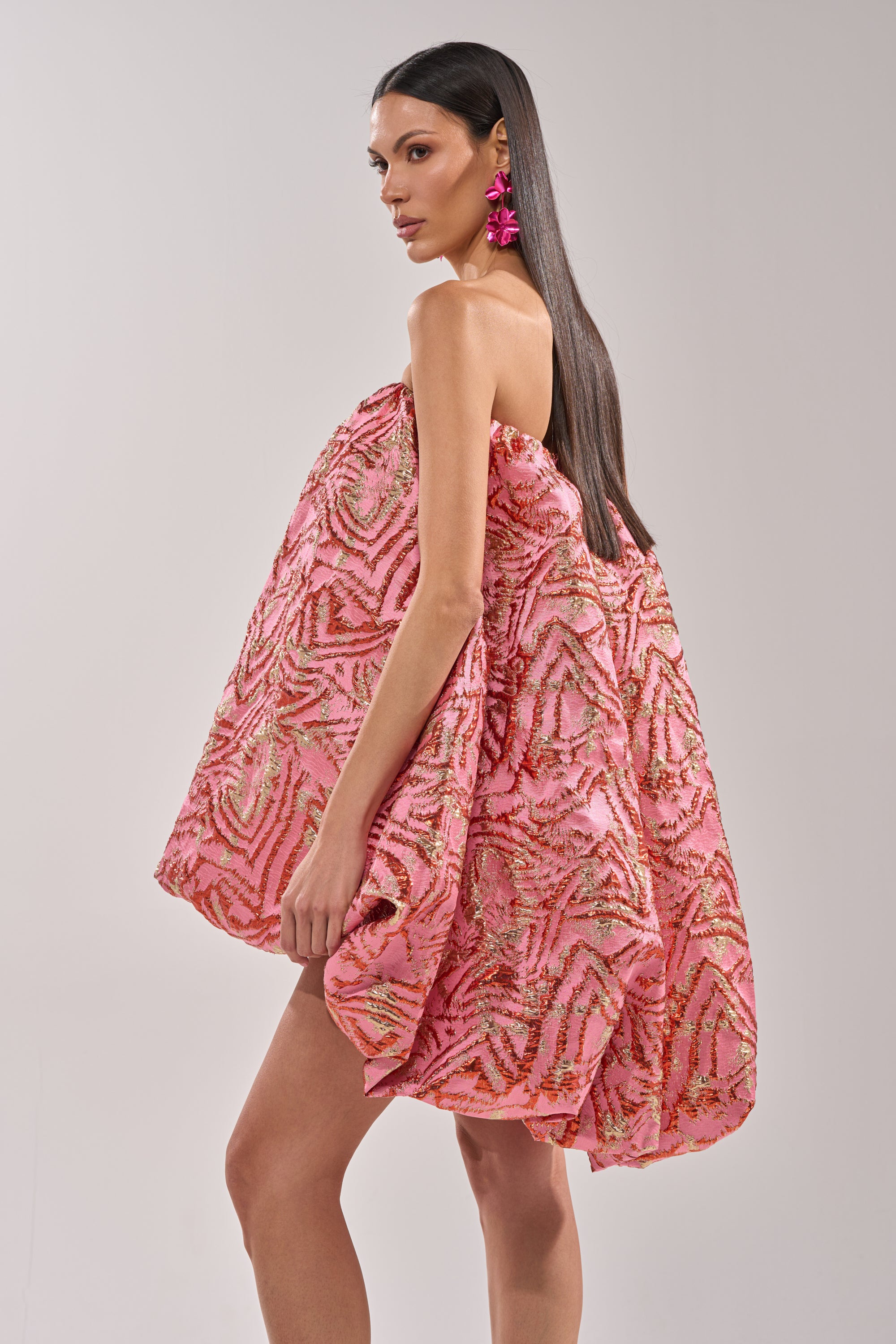 A woman stands in profile against a plain light background, wearing the LOVER GIRL BROCADE BUBBLE DRESS IN PINK with a geometric pattern and textured fabric. She has long straight hair and bold pink flower earrings. 2