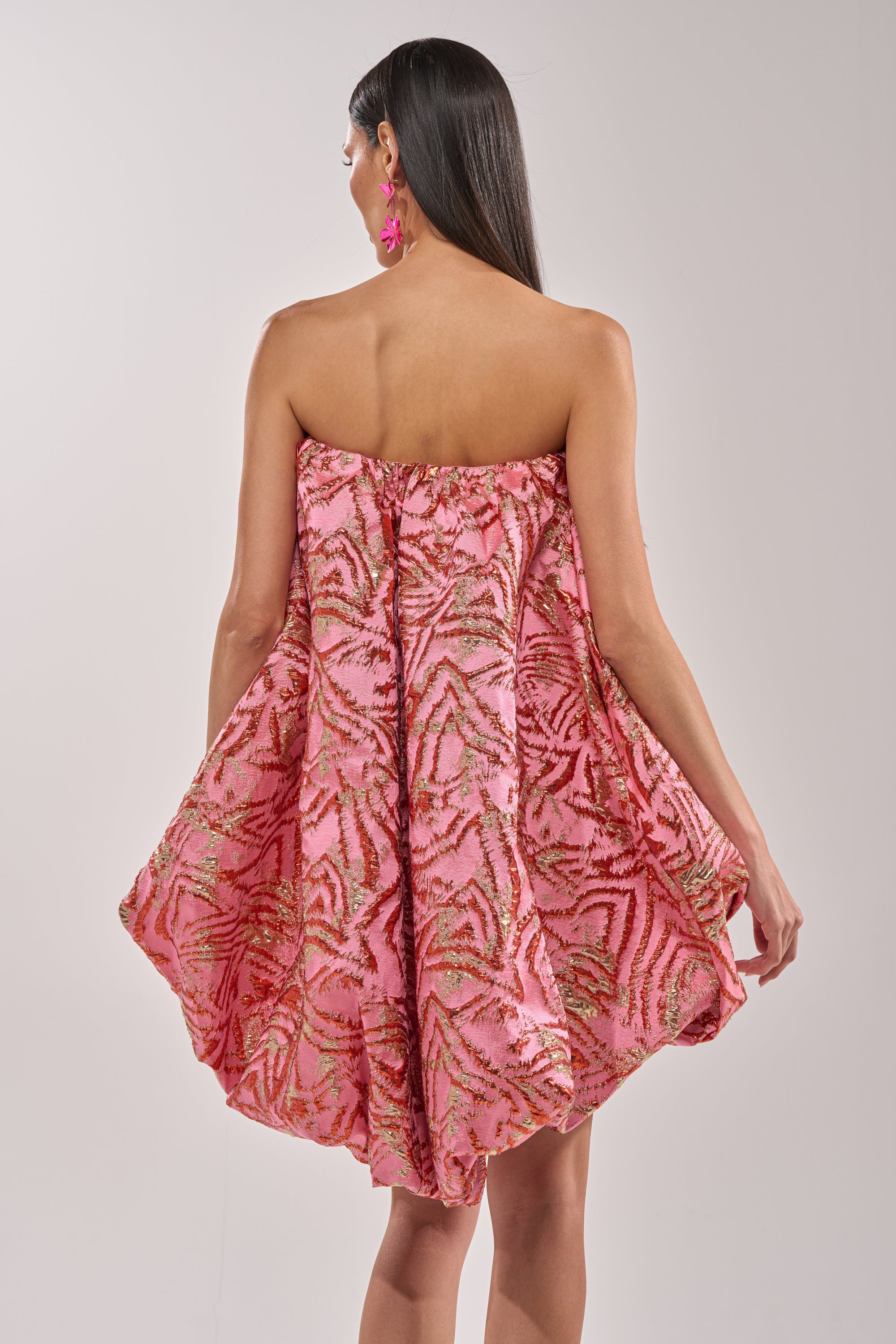 A woman with long dark hair faces away from the camera, showcasing the LOVER GIRL BROCADE BUBBLE DRESS IN PINK with its strapless design and gold and red patterns, highlighting the elegant and flowy brocade details. 3