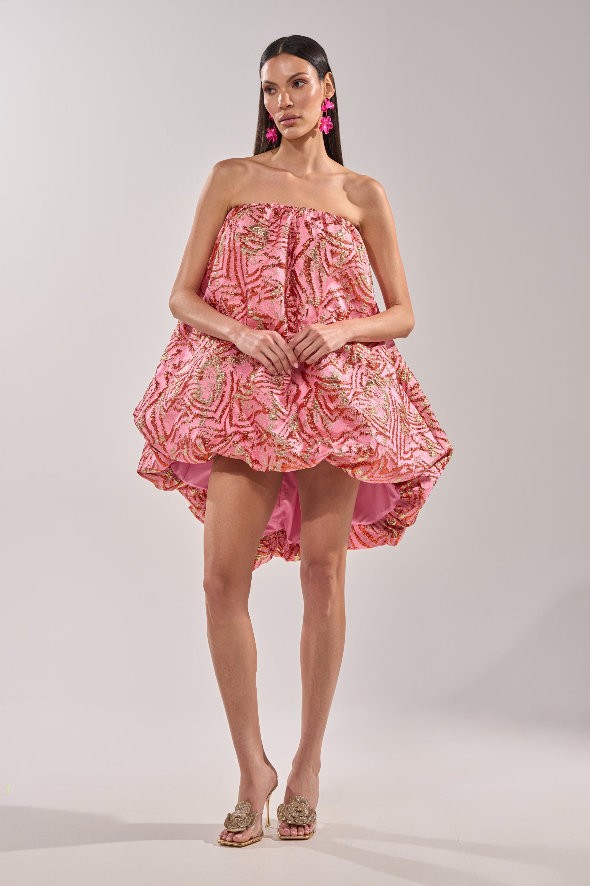 A woman wears the LOVER GIRL BROCADE BUBBLE DRESS IN PINK, a strapless mini with a pink and gold leaf pattern, styled with pink statement earrings and beige open-toe heels against a plain light background. 4