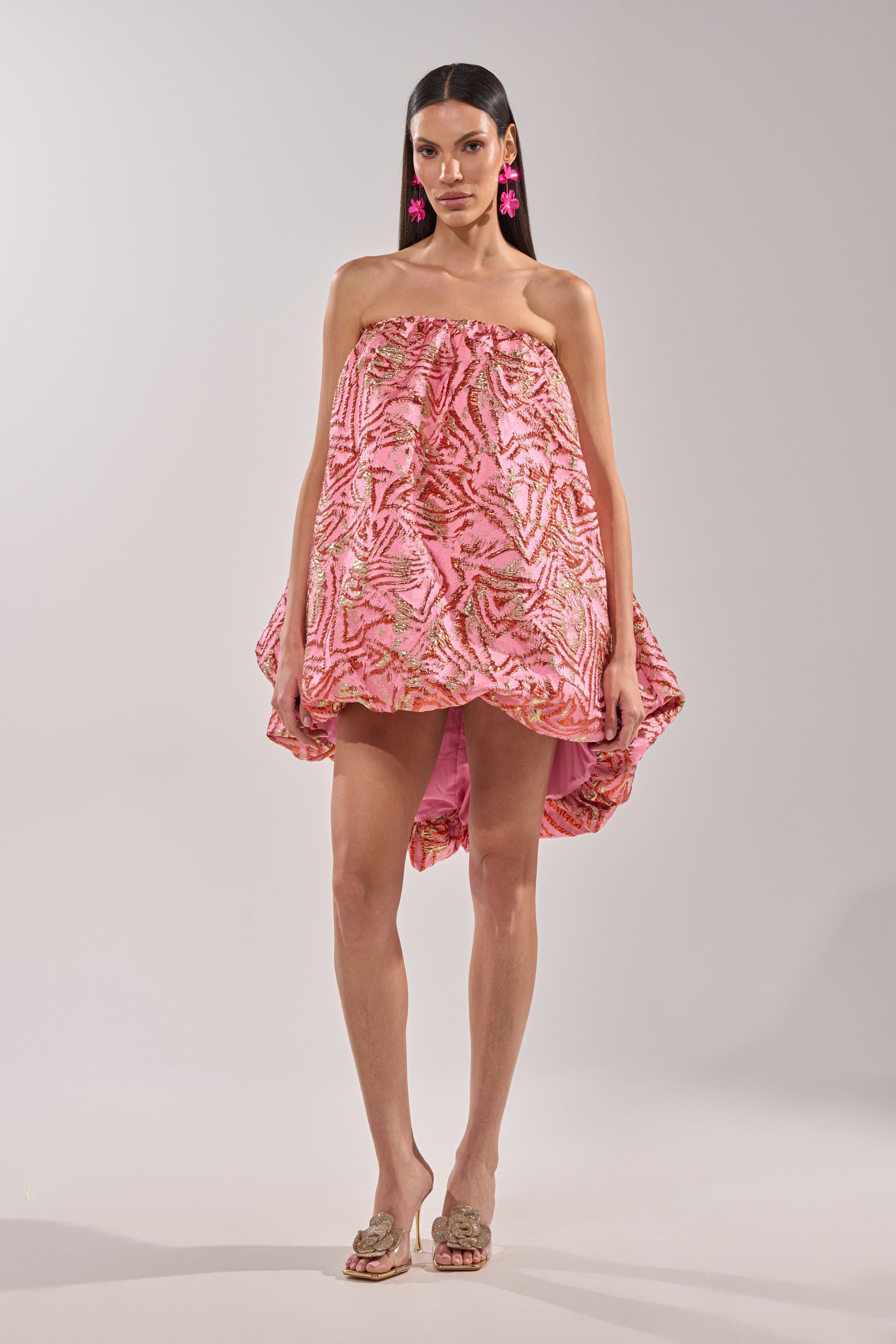 A woman stands against a plain background wearing the LOVER GIRL BROCADE BUBBLE DRESS IN PINK, a strapless mini dress with a pink and gold metallic finish, voluminous silhouette, and high-low hem. She styles it with pink floral earrings and beige open-toe heels. 5
