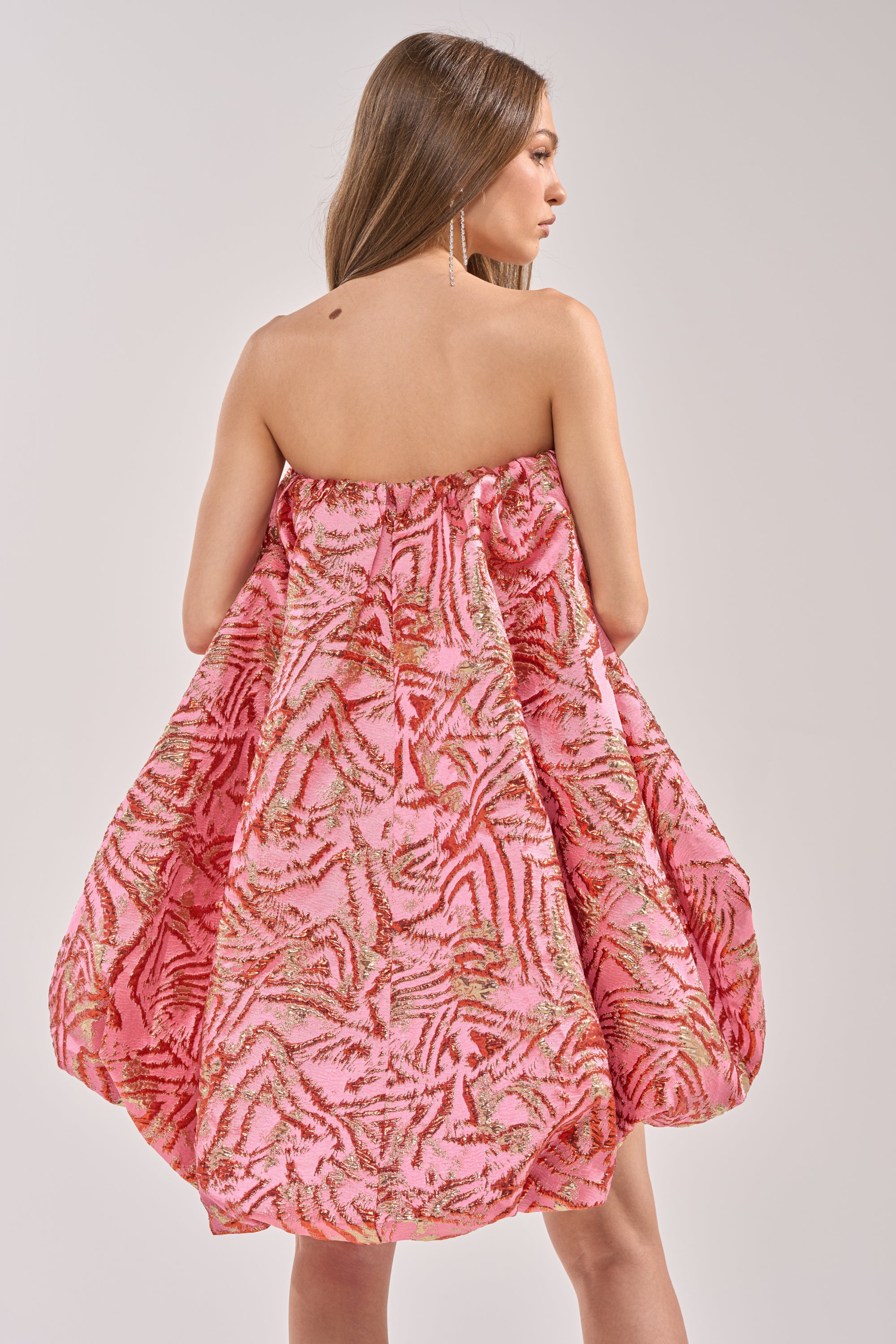 A woman with long brown hair models the LOVER GIRL BROCADE BUBBLE DRESS IN PINK, a strapless mini dress with red and gold abstract patterns, standing against a plain light background. 5
