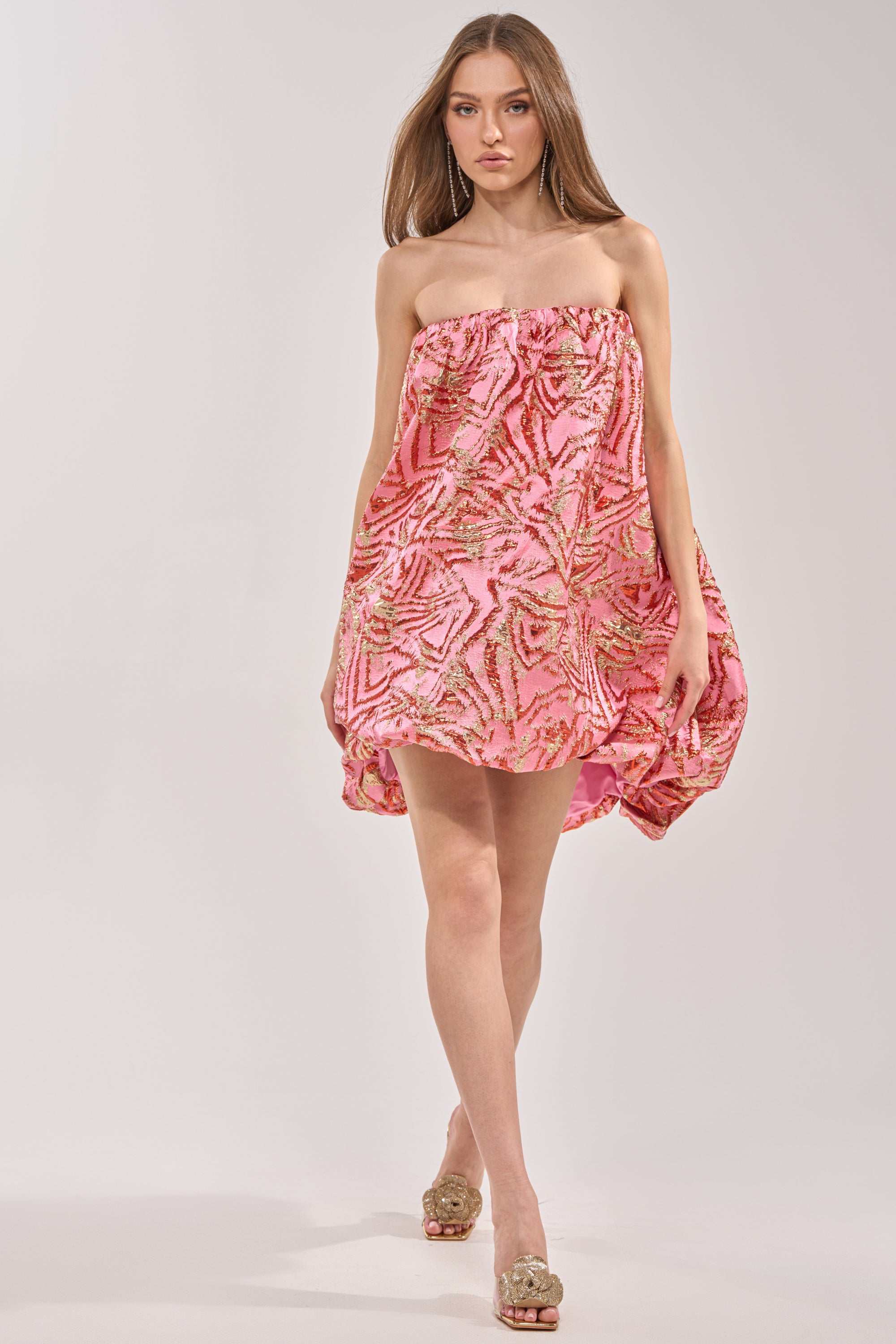 A woman walks confidently in the LOVER GIRL BROCADE BUBBLE DRESS IN PINK, a strapless mini with a voluminous silhouette. She pairs it with gold open-toe heels, her long straight hair flowing against a light neutral background. 6
