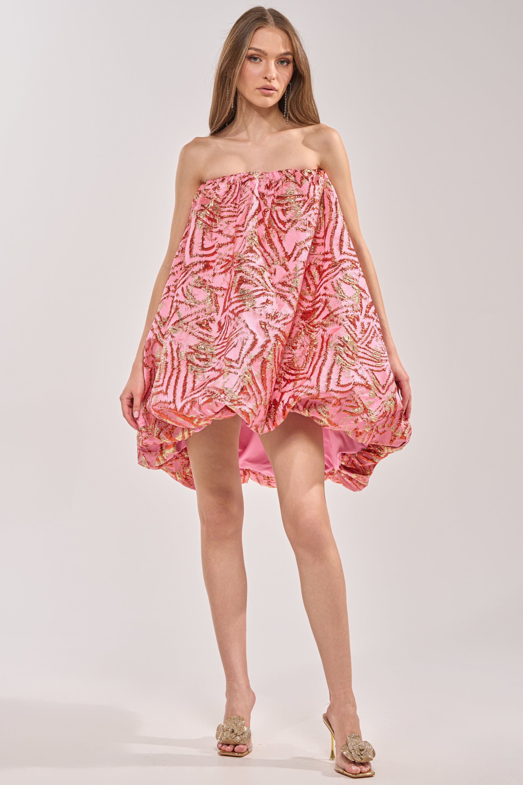 A woman wears the LOVER GIRL BROCADE BUBBLE DRESS IN PINK, a strapless mini featuring gold and red abstract patterns, a voluminous pink bubble silhouette, and a high-low hem. She pairs it with open-toe heels against a plain light background. 7